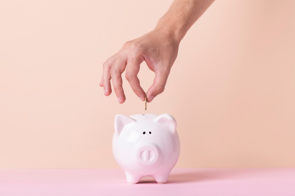 A hand ready to insert a coin into a pink piggy bank.